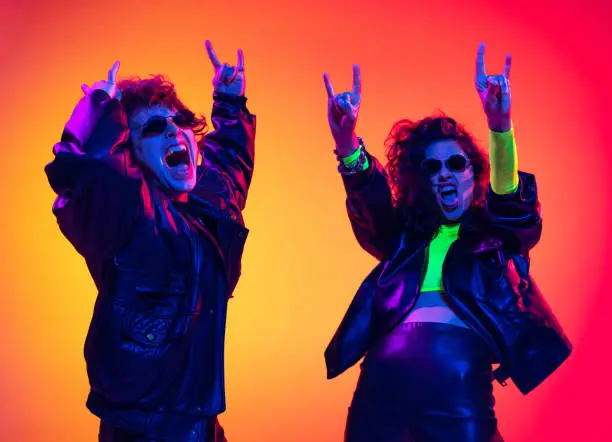 Rock music fans. Excited young man and girl in black leather outfits having fun, gesturing, shouting over yellow-red background in neon light. Concept of style, music, fashion and youth