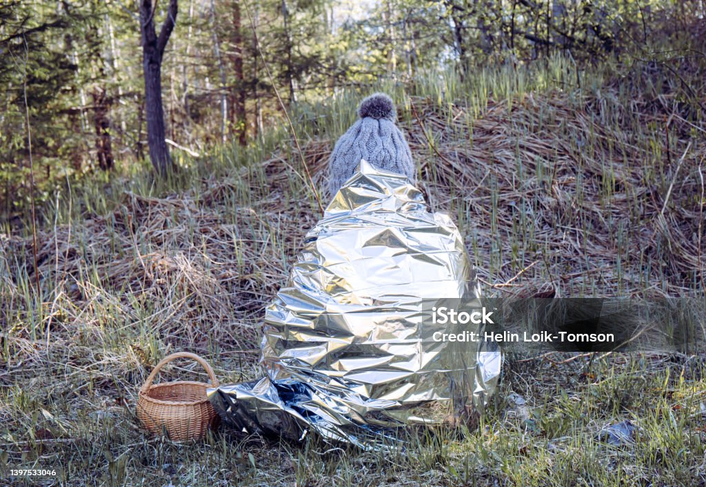 Woman person is lost and sit in wild forest in cold day, using first aid emergency blanket to prevent hypothermia and body heat loss. Emergency blanket concept. First Aid Stock Photo