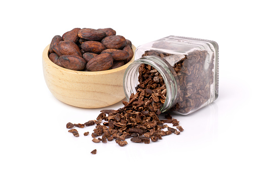 Cocoa nib in glass bottle with dry cacao beans in wooden bowl isolated on white background.