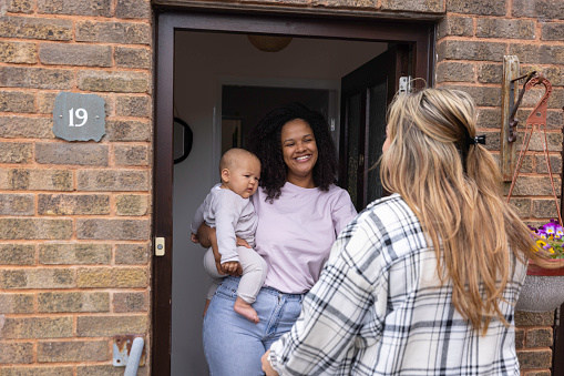 Multiracial mother holding her baby whilst standing in her doorway chatting to her friend.