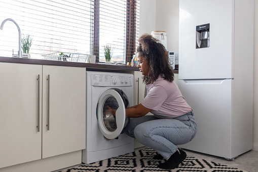 Single multiracial young adult kneeling down to put the dirty clothes into the washing machine.