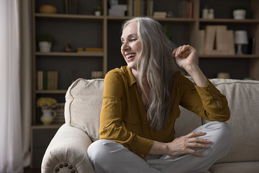 Cheerful beautiful old mature 50s woman sitting on couch at home, looking away, laughing, smiling at good thoughts, thinking, touching grey hair. Happy retirement, insurance concept