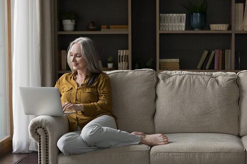 Happy senior laptop user woman sitting on couch at home, enjoying leisure time, chatting online, watching movie, shopping on Internet. Pretty freelance business lady working at computer. Full length