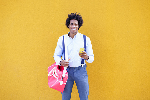Guy with curly hair wearing shirt and suspenders. Black man with afro hairstyle carrying a sports bag and smartphone against a yellow urban background.