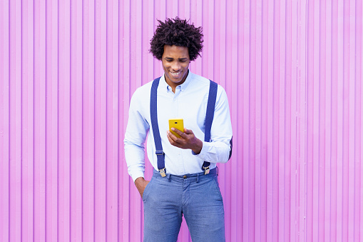 Black man with afro hairstyle using a smartphone against a pink blinds background. Guy with curly hair wearing shirt and suspenders.