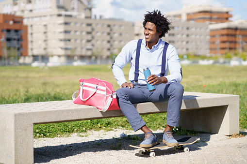 Black working man with afro hair taking a coffee break sitting on a park bench with his feet on his skateboard.