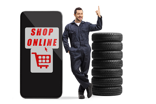 Full length portrait of an auto mechanic with tires next to a big smartphone with text shop online isolated on white background