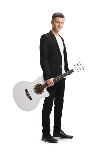 Elegant young man in a black suit holding an acoustic guitar and looking at camera isolated on white background