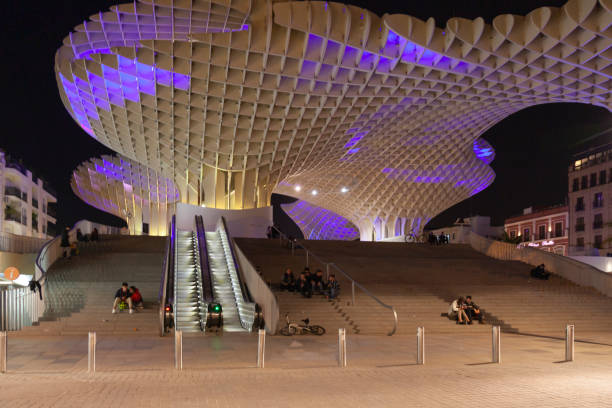 Seville, Spain - 7th April 2022. Young people sitting under Las Setas de Sevilla, aka Las Setas de la Encarnación, which is illuminated by coloured lights at night. The structure was designed by Jürgen Mayer H, built out of Finnish birch wood, and completed in 2011. N.B. Focus is on the parasols.