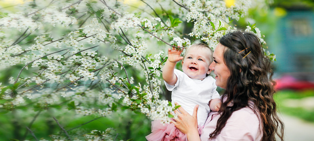 Lovely beautiful caucasian mom with her 1 year old daughter in spring blooming flowers. The concept of natural, conscious motherhood, female natural beauty and wavy hair.Young european woman with baby