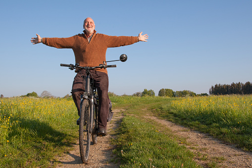 Fresh air, nature, freedom, bike. A senior citizen rides his bike hands-free on a dirt road through beautiful nature, laughing and stretching his face towards the sun.