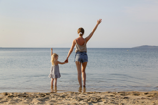 Mother and daughter on the beach looking to horizon over the sea with hands raised up. Family summer vacation concept.