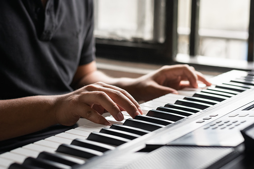 detailed views of hands playing a synthesizer. young man studying piano at home with lighting coming into his room through his window. concept of music production and artistry