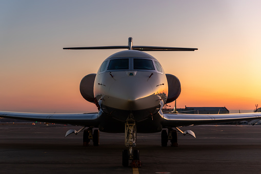 Scenic front view modern luxury expensive private jet plane parked airport taxiway hangar warm colorful dramatic evening warm sunset sun light sky background. Executive aicraft vip travel concept.