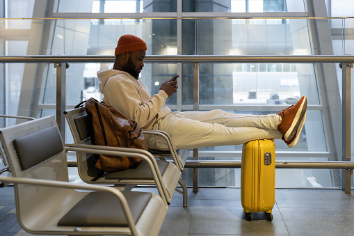 Pensive African man sitting in hall of airport terminal using mobile phone. Thoughtful passenger travelling with backpack, online chatting in social media in smartphone and waiting landing on board