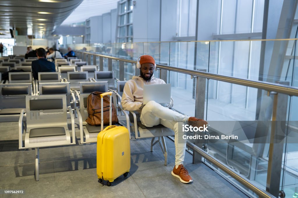 Happy hipster black man sitting in airport terminal using laptop wear headphones and listening music Smiling African American man sitting in hall of airport terminal using laptop, wear headphones and listening music. Passenger travelling with backpack, online working on computer and waiting landing. Airport Stock Photo