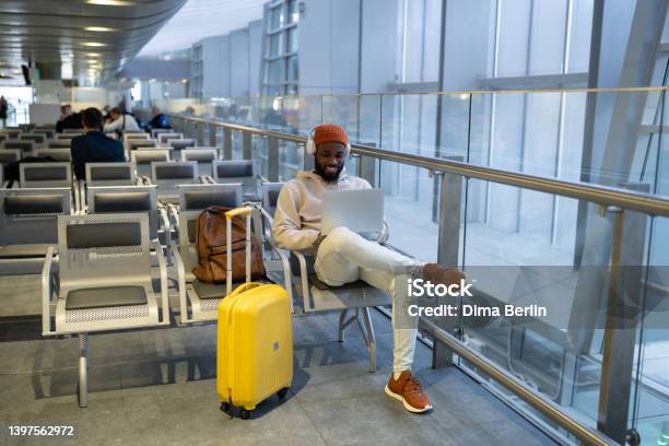 Happy Hipster Black Man Sitting In Airport Terminal Using Laptop Wear Headphones And Listening Music Stock Photo - Download Image Now