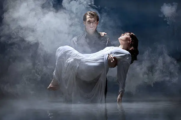 Dark haired man dressed all in black standing in pale, still lake water carrying a young maiden in white dress in a misty, midnight setting.