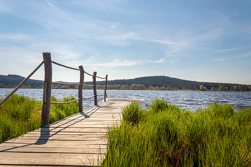 wooden pier with rope railing at Olsina pond, wooded hill and blue sky in the background, Czech Republic