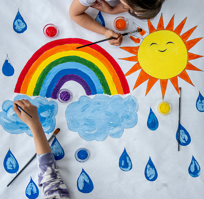 Two children are seen painting in this aerial photo.  They have a large white piece of paper laid out on the floor and are laying on their stomachs as they paint a mural of a sunshine, clods and rain together.