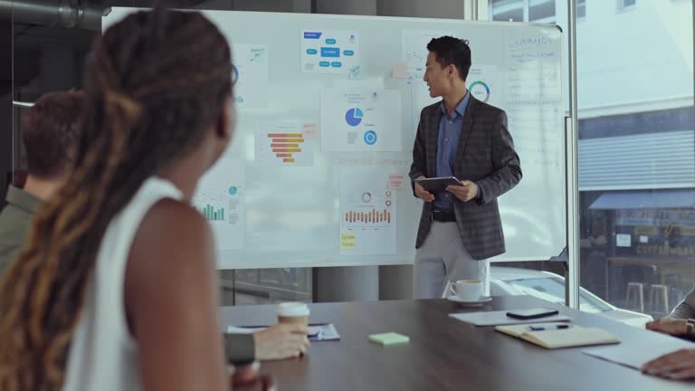 Young asian businessman doing a presentation at a whiteboard while holding a digital tablet with his colleagues in a boardroom at work. Chinese businessman talking at a whiteboard in a workshop at work