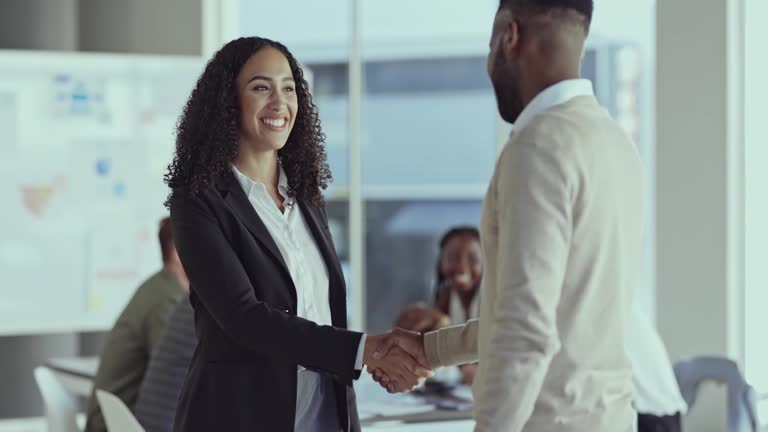 Young curly mixed race hispanic businesswoman smiling while greeting and shaking hands with an employee in an office at work. Two businesspeople shaking hands standing in an office at work