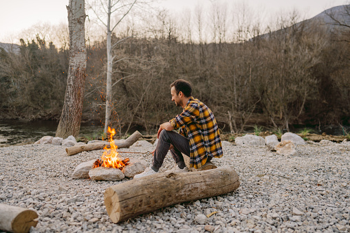 Photo of a young man sitting by the campfire and enjoying nature alone.