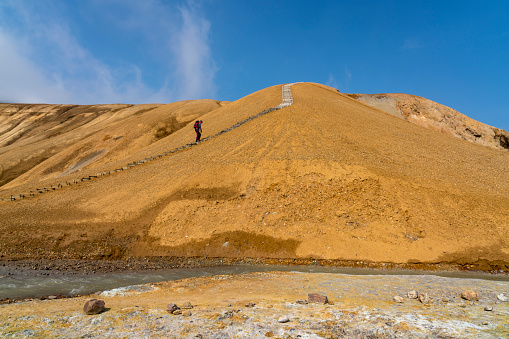 One tourist moving up the wooden stairs at barren volcanic landscape, Hveradalir geothermal area on Kerlingarfjoll mountain range against blue sky, Iceland