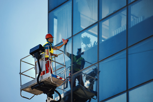 Side view of a mid 30's man washing windows on a residential building facade. He's safely standing inside construction platform.