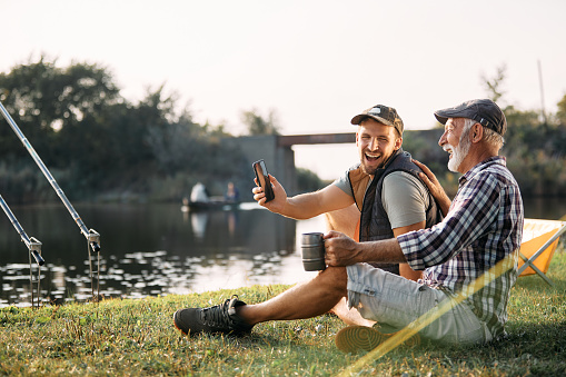 Happy man using smart phone and laughing with his senior father while fishing and camping in nature.