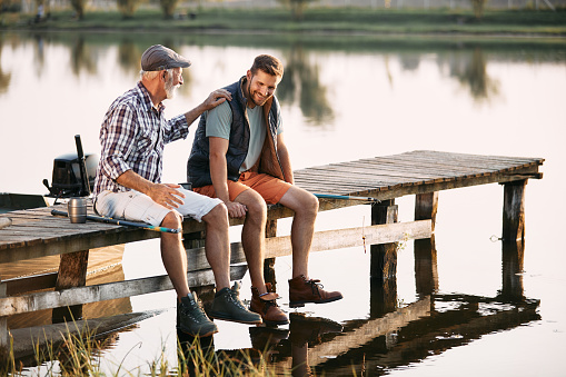 Happy mature man and his adult son communicating while relaxing on a pier and fishing by the lake. Copy space.