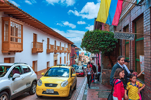 Bogotá, Colombia - July 20, 2016: One of the narrow streets in the historical La Candelaria district, in the South American capital city of Bogota. Some local Colombian people and traffic can be seen. The image was shot on 20th of July which is National Day in Colombia; the streets are therefore not as busy as they can normally be. It is in this area that the Spanish Conquistador, Gonzalo Jiménez de Quesada founded the city in 1538. The area is known for its historical Spanish colonial architecture; there are buildings in La Candelaria that go back a few humdred years. Many of the walls in the area are painted in the vibrant colours of Colombia. The altitude at street level is around 8,660 feet above mean sea level. Photo shot on a cloudy morning; horizontal format. Copy space.