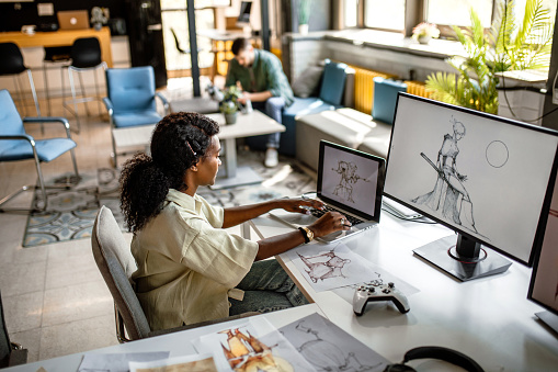 Young, black, confident video game designer in company studio, working at her desk computer, drawing new sketches.