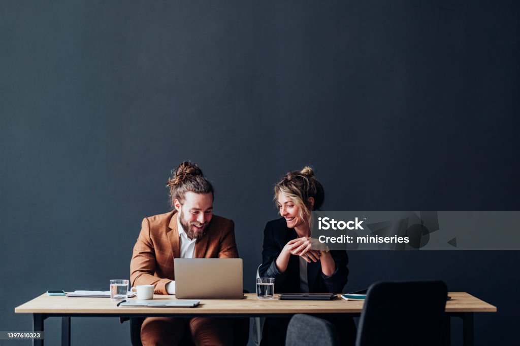 Two Smiling Colleagues Working Together On A New Project Happy businessman and businesswoman using a laptop computer while talking and sitting together at office desk. Office Stock Photo