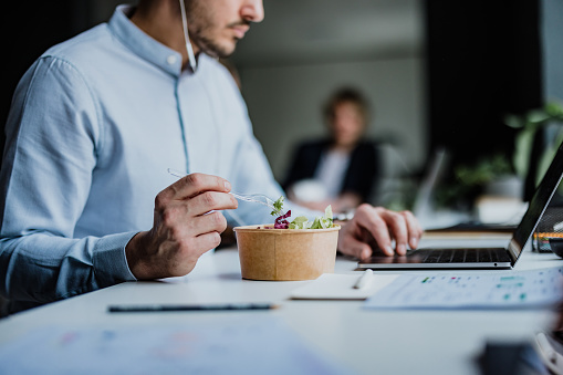 Unrecognizable businessman with earphones speaking on a conference call using a laptop computer while eating a healthy fresh salad in the modern office.