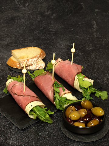 Pastrami wrapped with Swiss cheese and arugula and placed on a black slate plate.