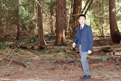 A high school student is walking in the forest. He is looking at the camera and smiling. He is wearing school uniform with suit, vest and tie.