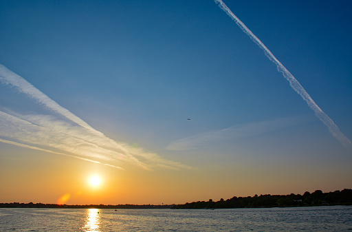 Vapor trails slash a blue sky as a plane flies over a lake at sunset.