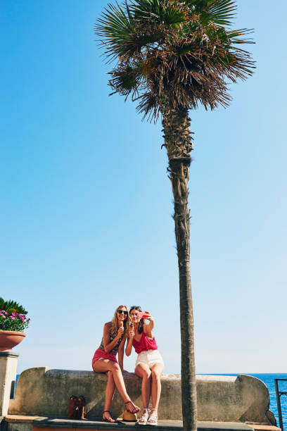 due amici che scattano selfie mangiando il gelato sulla spiaggia usando lo smartphone per connettersi in viaggio d'avventura - sea coastline europe tourist foto e immagini stock