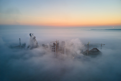 Aerial view of cement factory with high concrete plant structure and tower crane at industrial production site on foggy morning. Manufacture and global industry concept.