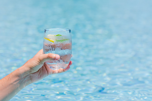 Hand holding a glass of cold drinking water with a piece of lemon on blue color background of swimmin pool.