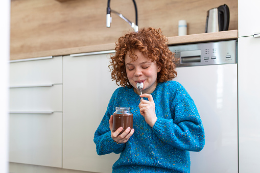 Young woman eating chocolate from a jar while sitting on the kitchen floor.