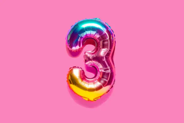 Rainbow foil balloon number, digit three. Top view. Colored numeral on pink background.