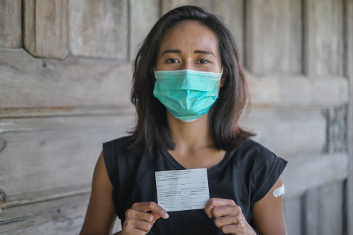 Close-up shot of Indonesian woman with surgical mask showing her COVID-19 vaccination record card