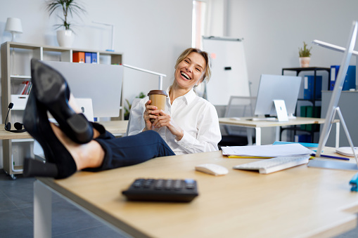 Joyful business woman with a cup of coffee putting legs on the table in office
