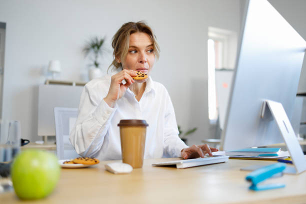 空腹のビジネスウーマンオフィスワーカーがコンピューターで作業しながらクッキーを食べている - chocolate candy unhealthy eating eating food and drink ストックフォトと画像