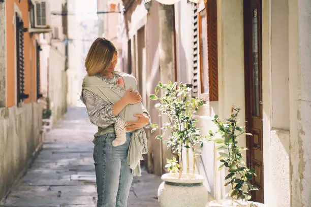 Photo of Babywearing. Mother and baby walking in town. Baby in wrap carrier. Young woman carrying little child in baby sling in mint color. Concept of green parenting, natural motherhood, postpartum period.