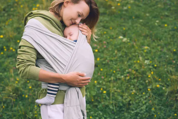 Photo of Babywearing. Mother and baby on nature outdoors. Baby in wrap carrier. Woman carrying little child in baby sling in green mint color. Concept of green parenting, natural motherhood, postpartum period.