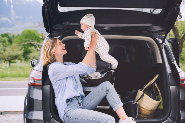 Mother with baby sitting in open car trunk outdoors. Road trip with baby. Travel in Europe. Young mother with her little baby having picnic near by the car. Safety driving by car with small kids. madre con el bebé sentado en el maletero abierto del coche al aire libre. viaje por carretera con bebé. viajes por europa. joven madre con su pequeño bebé haciendo un picnic cerca del coche. conducción segura en coche con niños pequeños.
