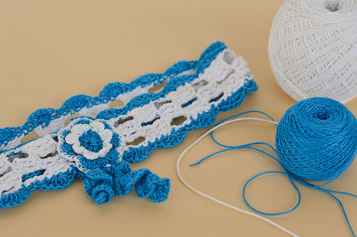 A hair band made of white and blue cotton threads crocheted, threads and a crochet hook on a beige background, diy.